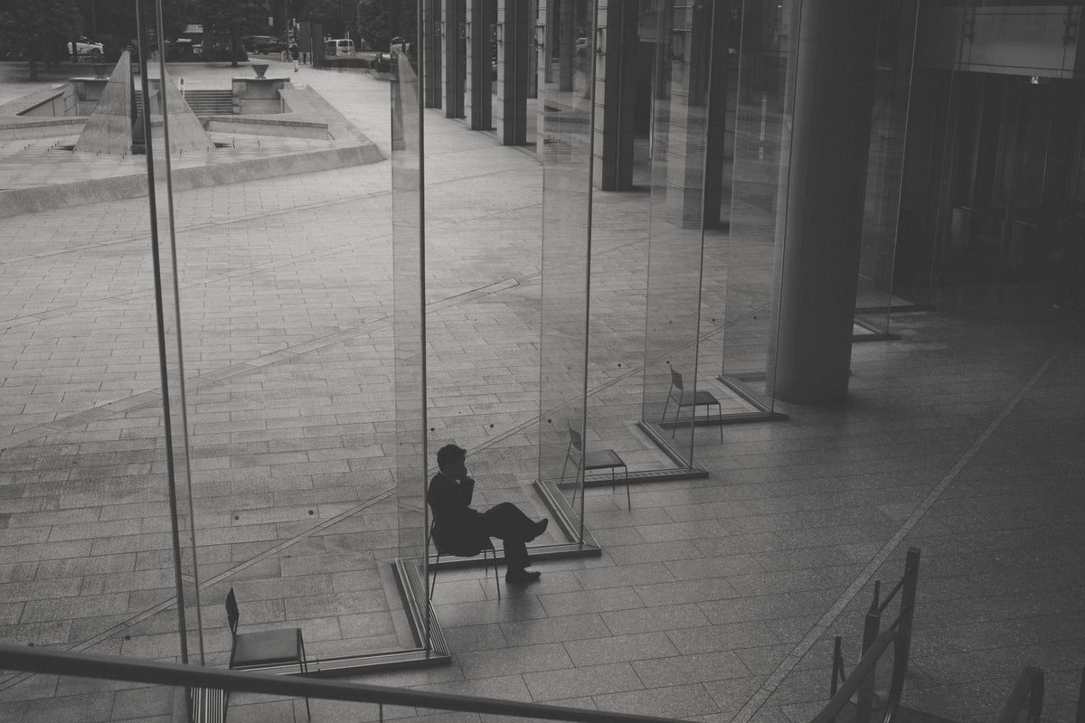 A man sitting alone among glass walls in a plaza, black and white